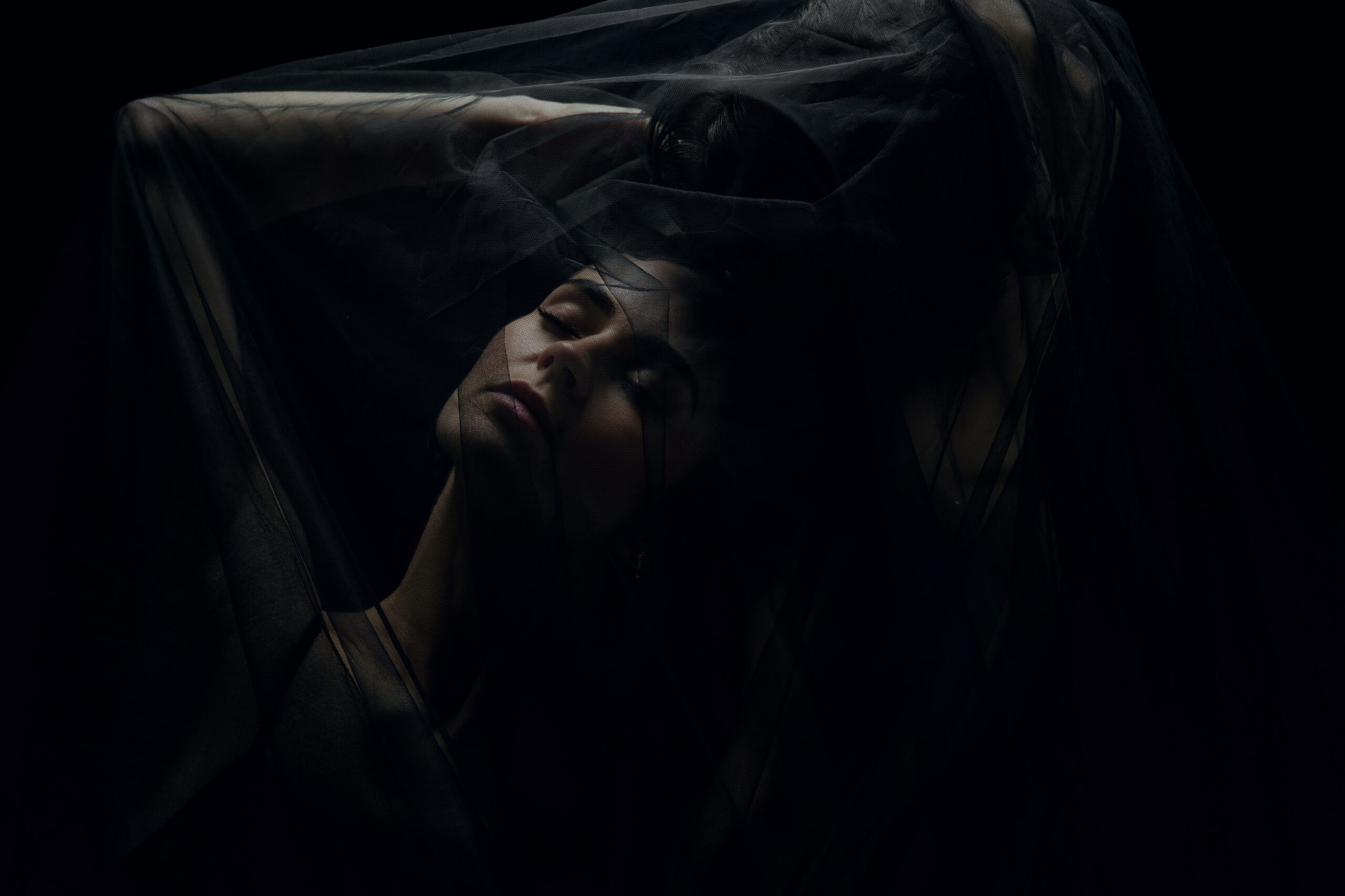 A woman's face tilted back beneath dark, sheer fabric, her eyes closed